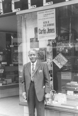 Curtis Jones in front of a radio/TV shop in Strasbourg, France, 1962. The poster in the window announces a Curtis Jones performance 'Recital de Blues Authentique' on June 28, 1962, organized by 'Le Club des Amis du Jazz' at 'Salle de l'Aubette' in Strasbourg, France; Courtesy of Pierre Monnery, who found a Curtis Jones LP with - amongst others - this photo inside; photographer's name not known